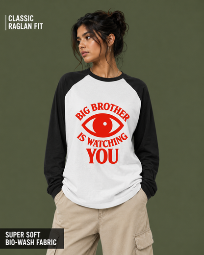 A woman wearing a raglan-style long-sleeve shirt with a white body and black sleeves, featuring a red graphic of an eye and the text 'BIG BROTHER IS WATCHING YOU' on the front. The background is a solid dark olive green.